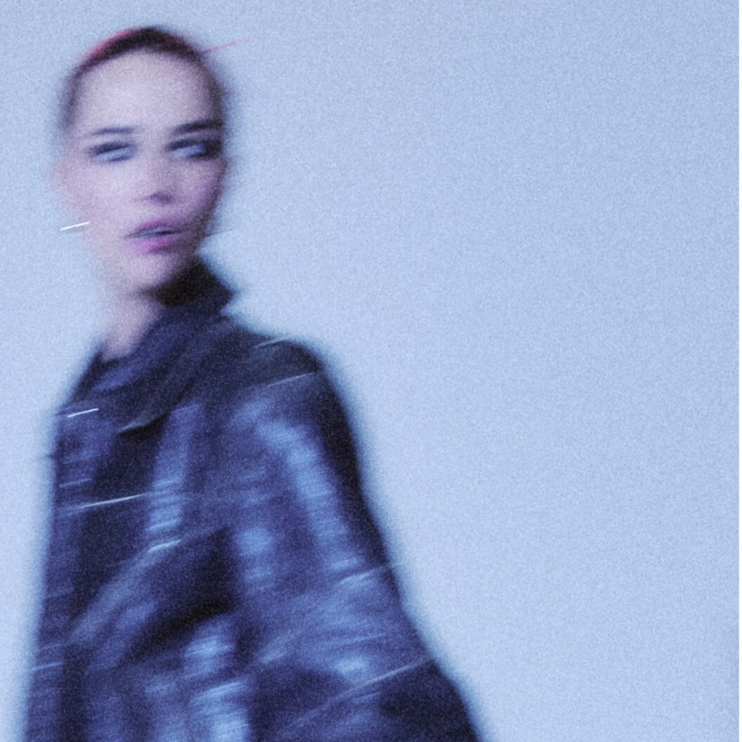 A waist-up grainy photo of a woman thinking about top unisex fragrance brand NOYZ while wearing a leather jacket walking quickly with a motion blur