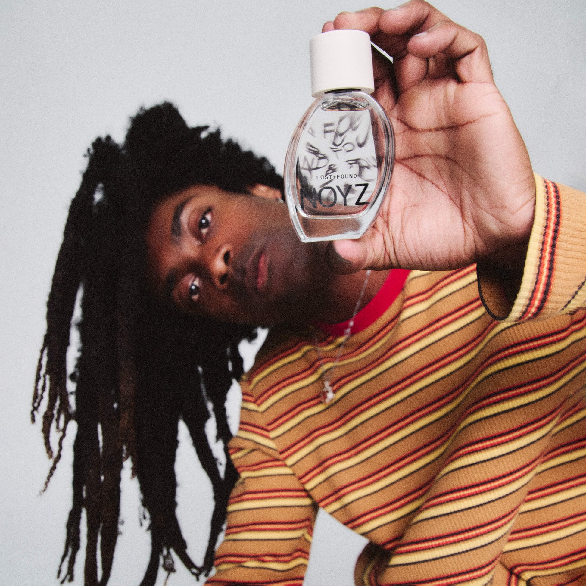 A photo of a man with dreadlocks holding a bottle of Noyz Lost + Found unisex fragrance