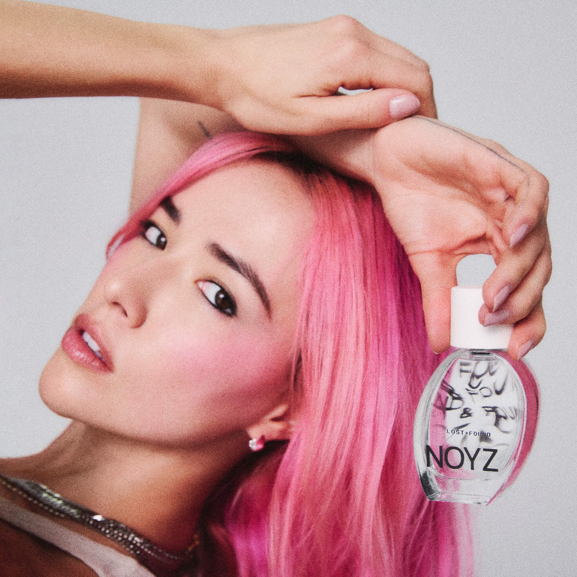 Close up photo of a woman with pink hair holding a bottle of Lost + Found by NOYZ fragrance.