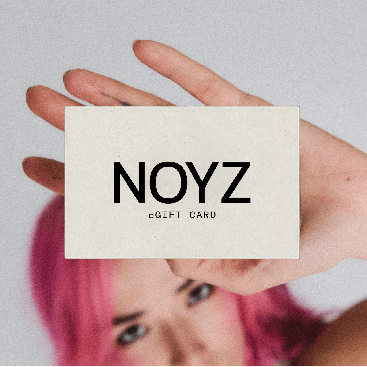 A unisex fragrance brand NOYZ gift card sits on top of a photo of a woman with pink hair and tattoos