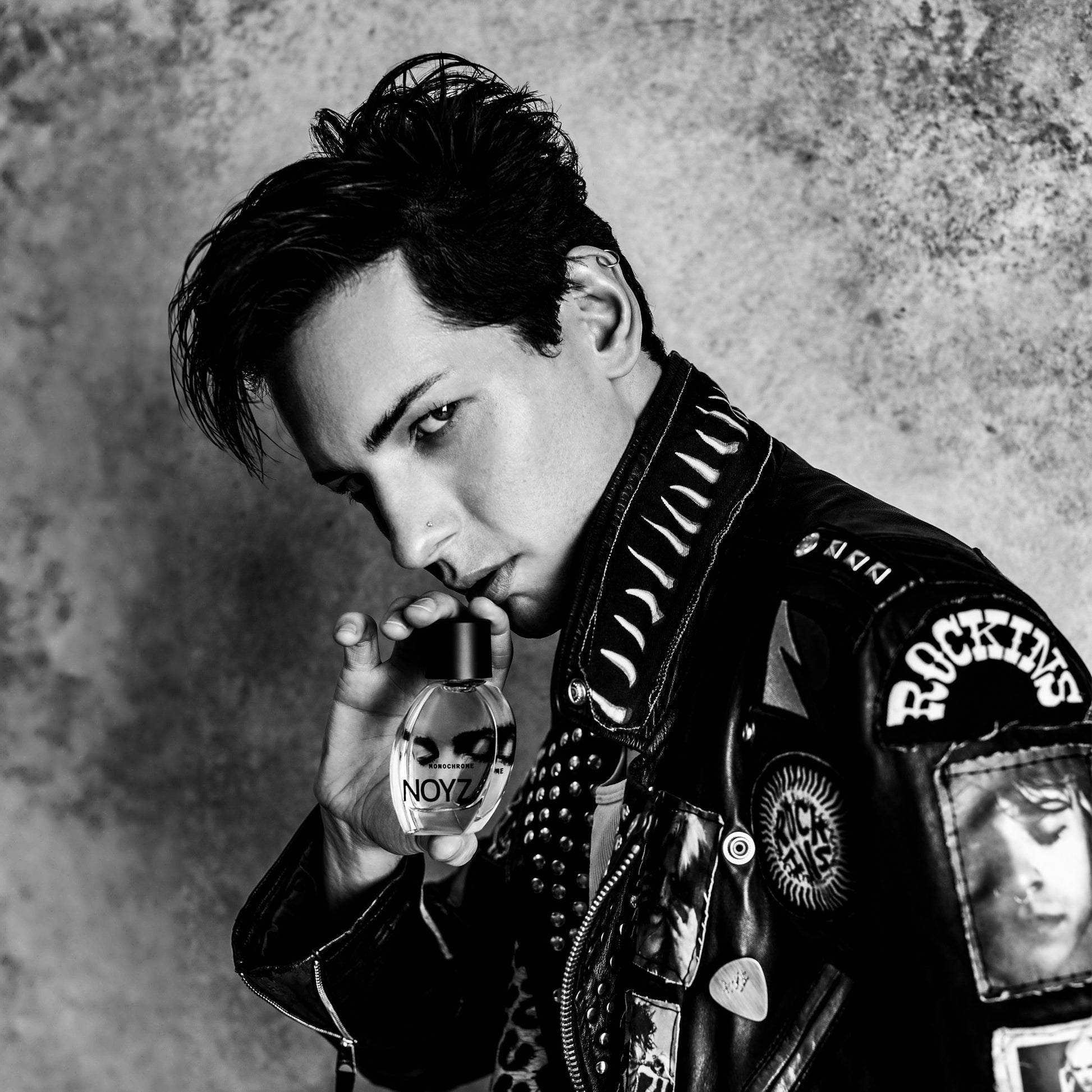 A man wearing a leather jacket with patches holding NOYZ Monochrome fragrance by Damon Baker.