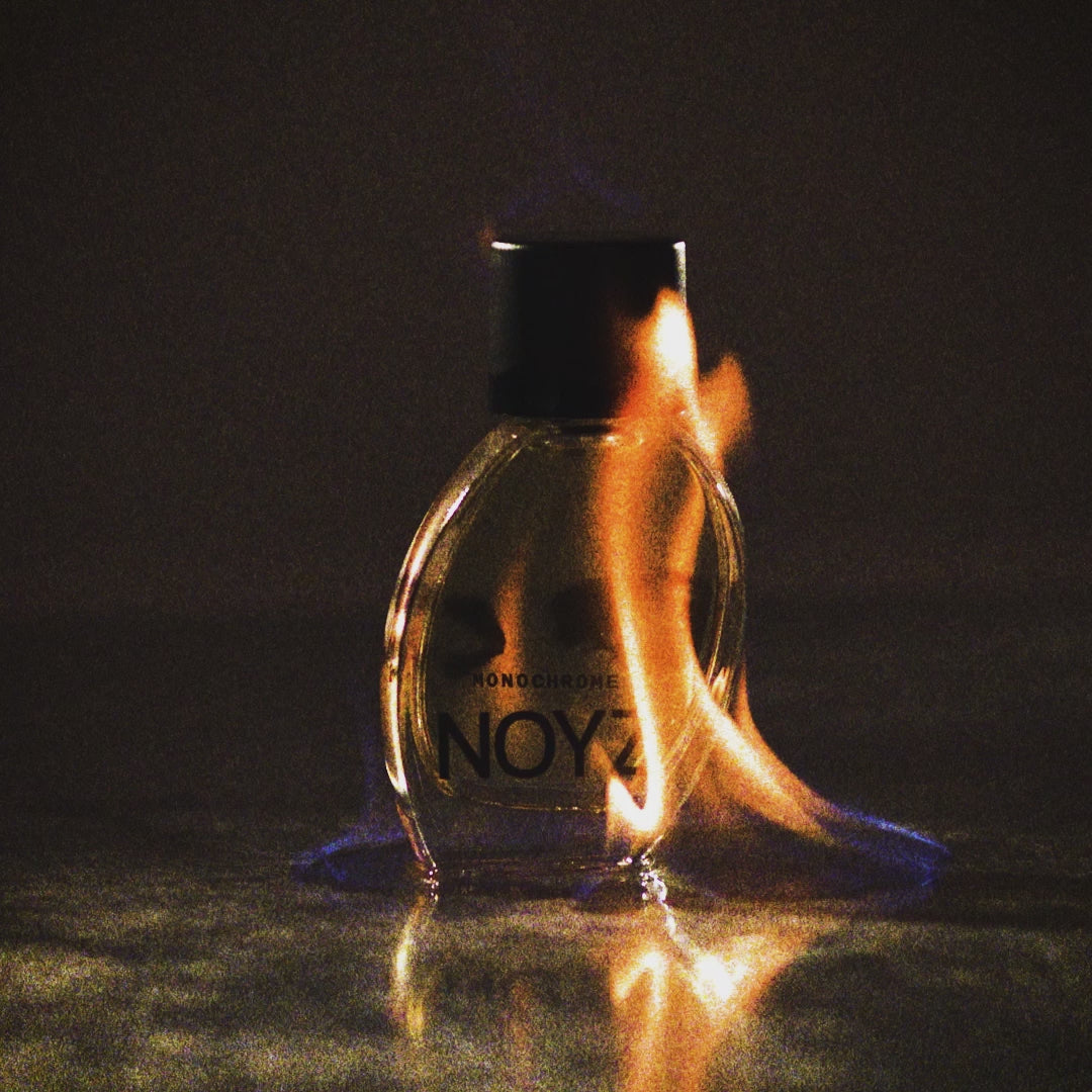 A bottle of Monochrome unisex perfume by NOYZ with a flame behind it in a dark and moody environment 